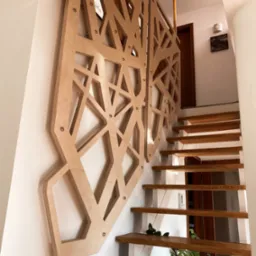 Ażurowa balustrada schodowa wykonana ze sklejki w geometryczne wzory, mocowana do ściany za pomocą metalowych śrub, z widocznymi drewnianymi stopniami schodów i rośliną doniczkową w tle.