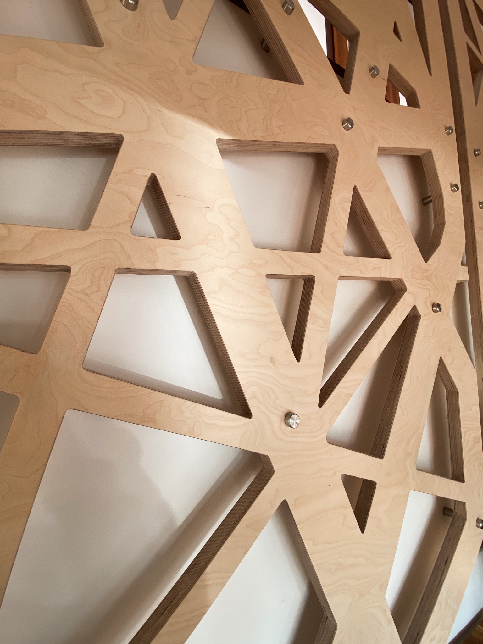 Ażurowa konstrukcja ze sklejki, przymocowana do ściany za pomocą metalowych śrub, tworząca geometryczny wzór z trójkątnych i trapezoidalnych otworów.