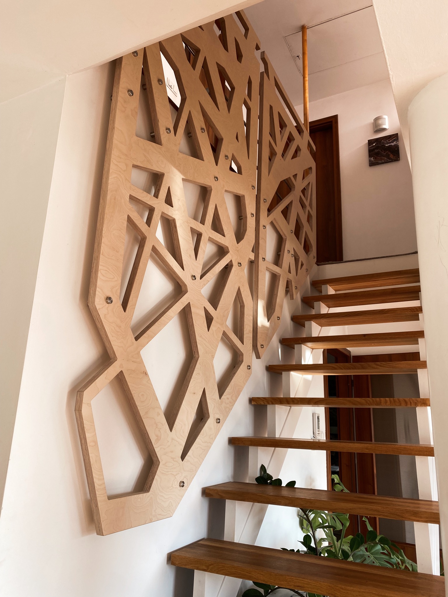 Ażurowa balustrada schodowa wykonana ze sklejki w geometryczne wzory, mocowana do ściany za pomocą metalowych śrub, z widocznymi drewnianymi stopniami schodów i rośliną doniczkową w tle.