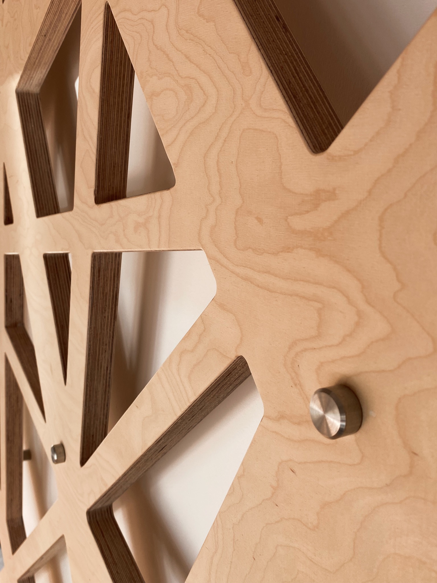 Ażurowa dekoracja ścienna ze sklejki drewnianej, mocowana do białej ściany za pomocą metalowych kołków dystansowych, widoczna faktura drewna i warstwowa struktura sklejki.