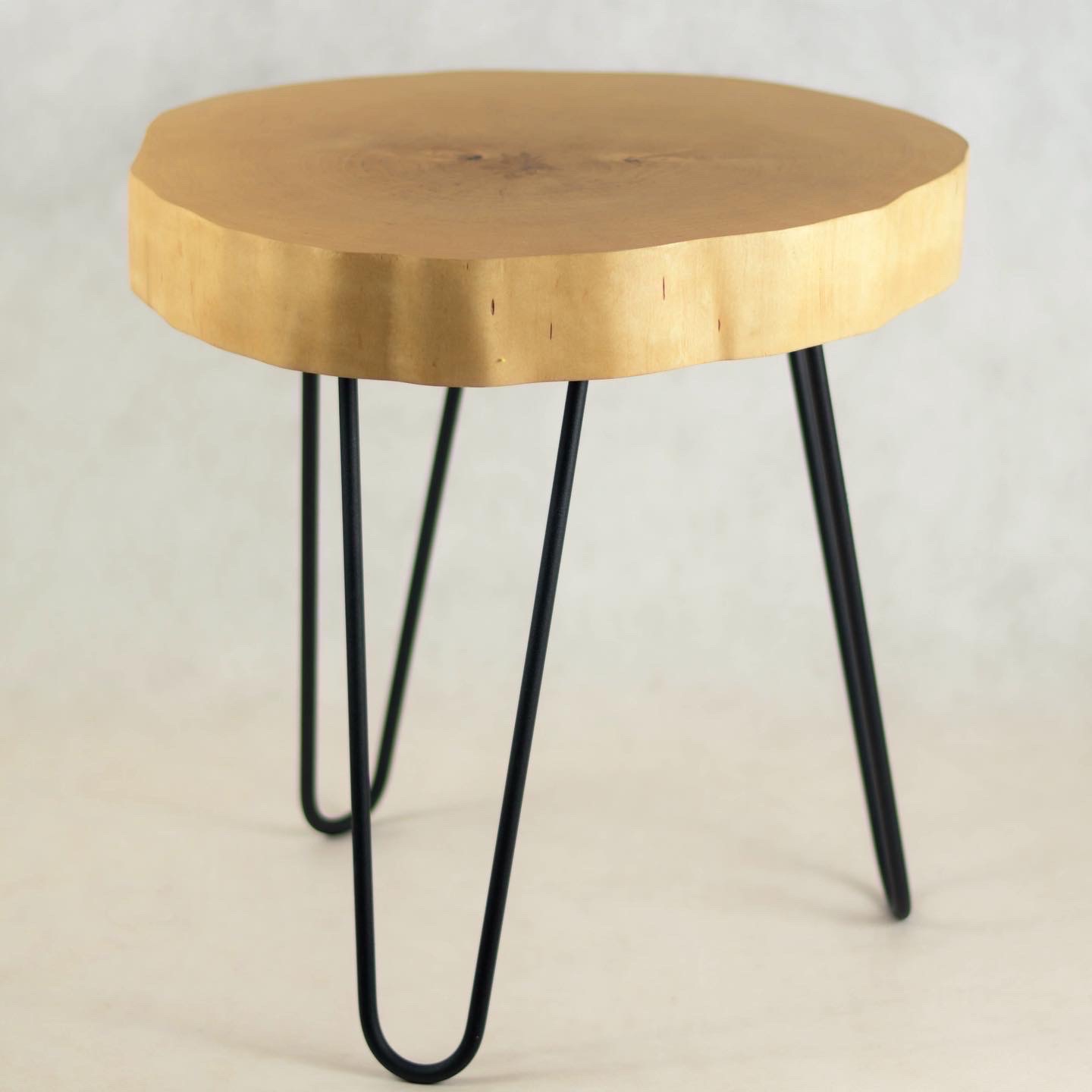 Mały, okrągły stolik kawowy z blatem z naturalnego drewna i czarnymi, metalowymi nogami w stylu hairpin, na jasnym tle.