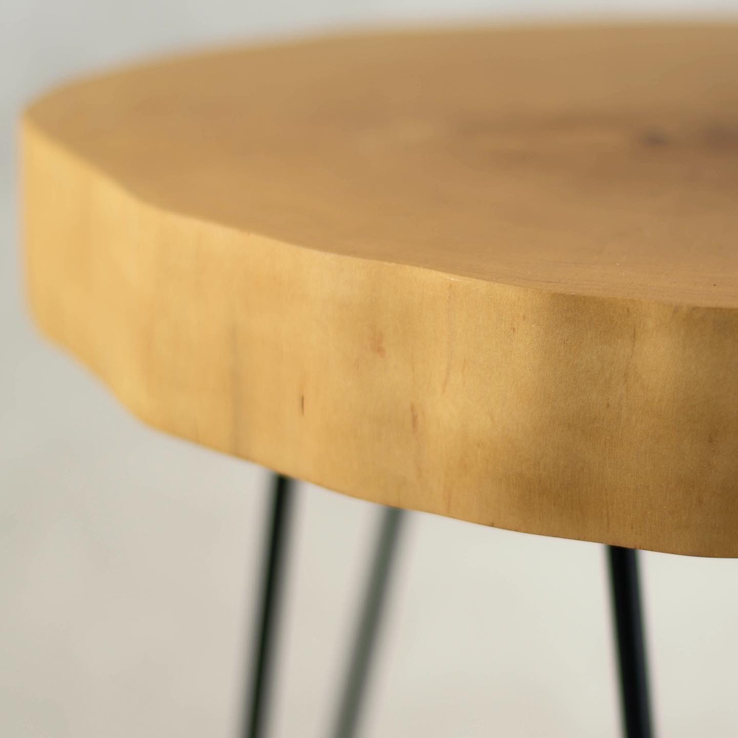 Okrągły blat z jasnego drewna z naturalnym obrzeżem, wsparty na trzech smukłych, czarnych metalowych nogach.