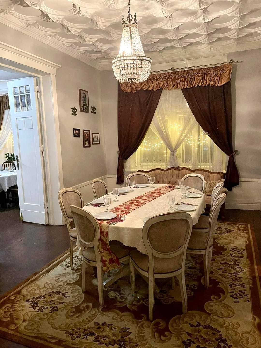 Elegancka sala jadalna z owalnym stołem nakrytym białym obrusem z czerwonym bieżnikiem, ustawiona pod kryształowym żyrandolem i ozdobnym sufitem, widoczne krzesła w stylu vintage, okno z brązowymi...