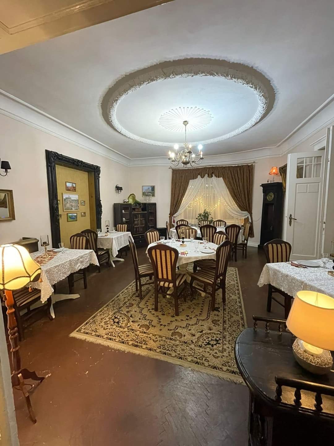 Elegancka sala bankietowa z okrągłymi stołami nakrytymi białymi obrusami, krzesła w paski, dywan w orientalne wzory, oświetlenie lampami i żyrandolem, dekoracja sufitu w stylu retro.