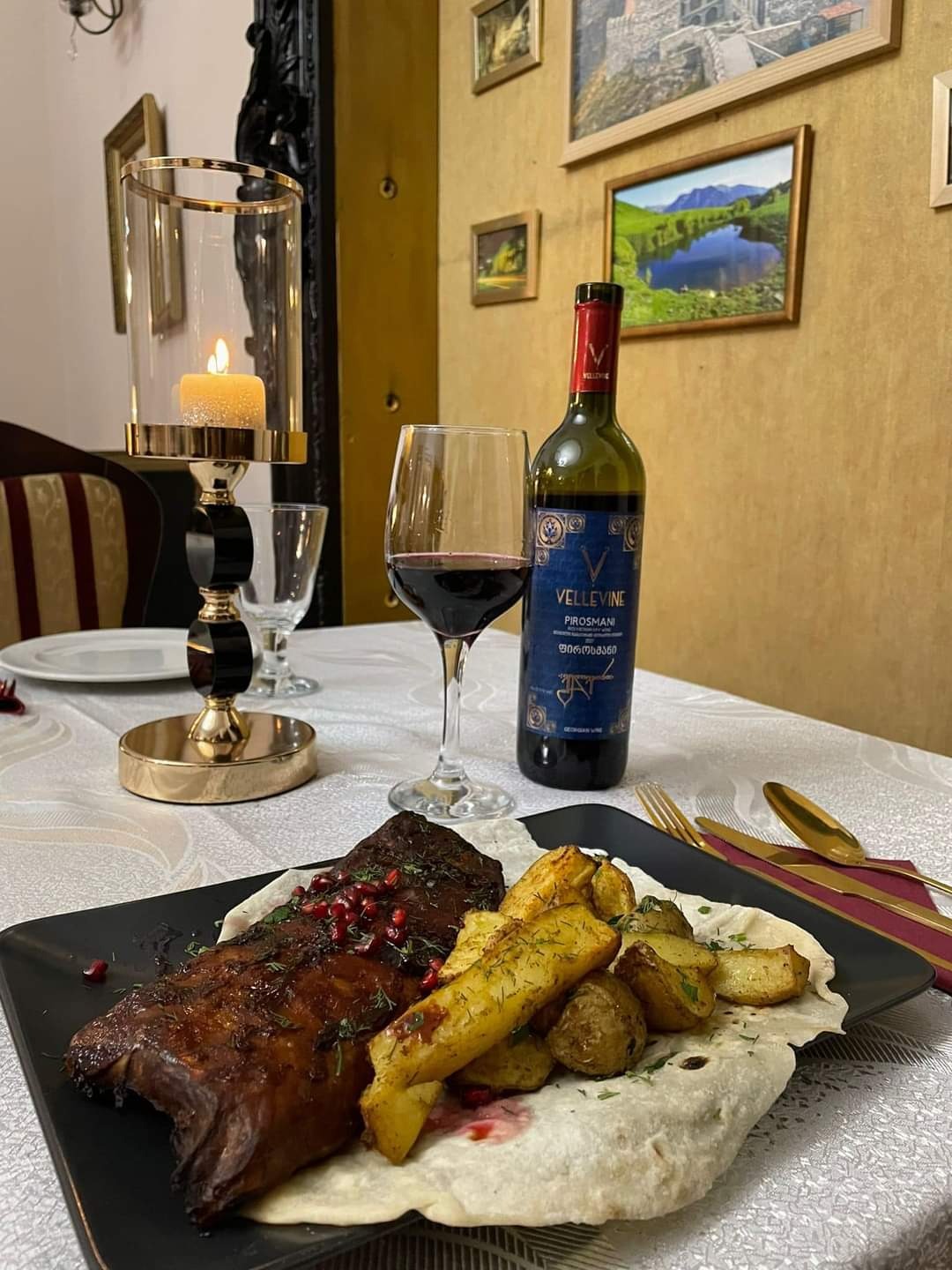 Uroczysta kolacja: pieczone mięso z granatem i ziemniakami na chlebku pita, wino VelleVine Pirosmani, świeca w złotym świeczniku, złote sztućce, obrazy na ścianie.
