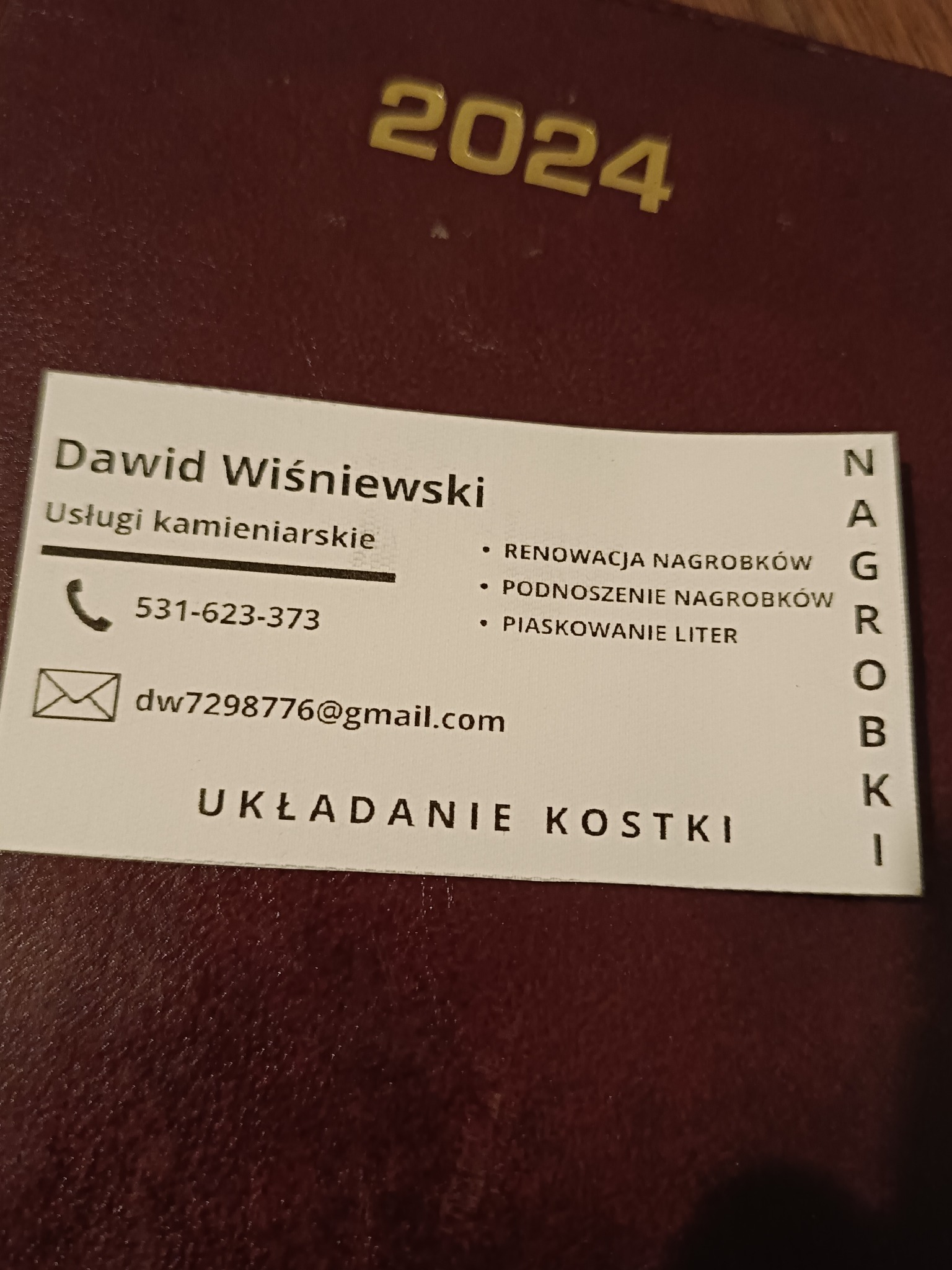 Wizytówka firmy kamieniarskiej Dawida Wiśniewskiego z numerem telefonu, adresem email, oferująca usługi renowacji, podnoszenia nagrobków, piaskowania liter oraz układania kostki, leżąca...
