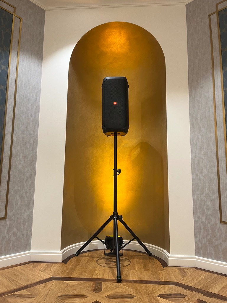 Czarny głośnik JBL na statywie, oświetlony od dołu, umieszczony we wnęce ze złotym tłem, na parkiecie.