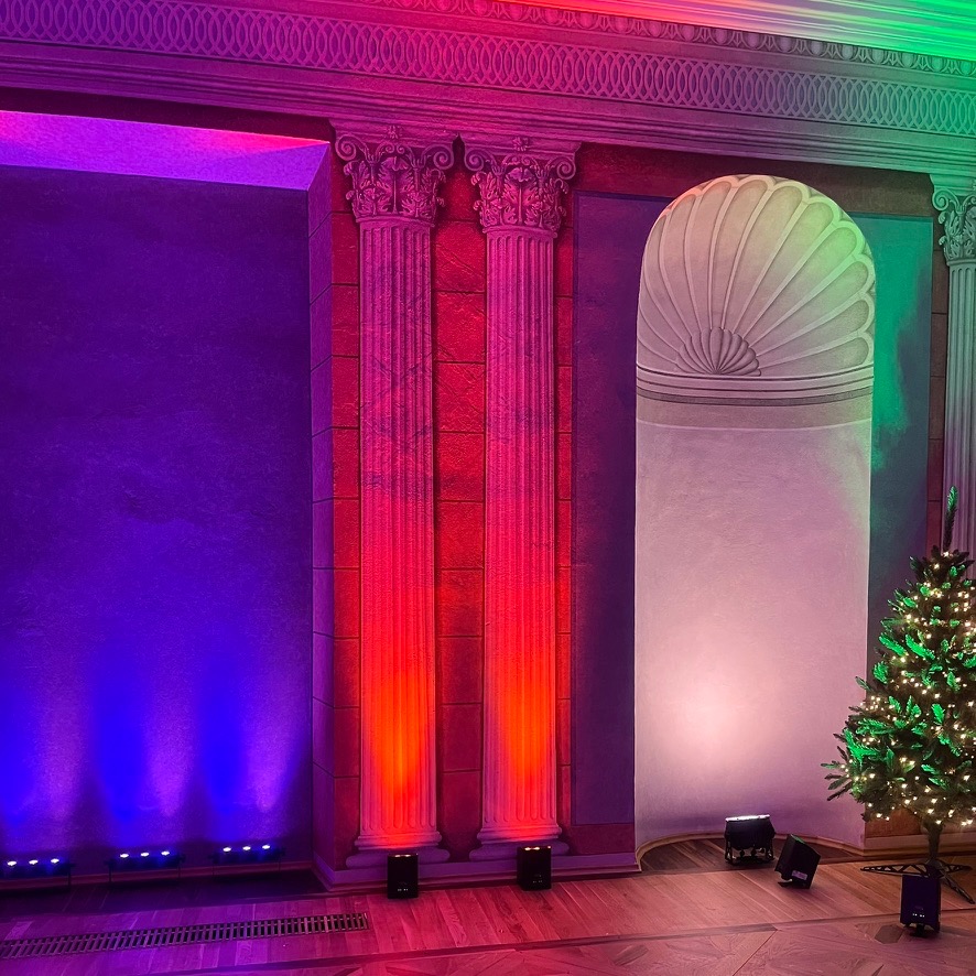 Dekoracja eventowa z kolumnami podświetlonymi na czerwono, ścianą podświetloną na niebiesko i zielono oraz choinką w rogu, oświetlenie sceniczne na podłodze