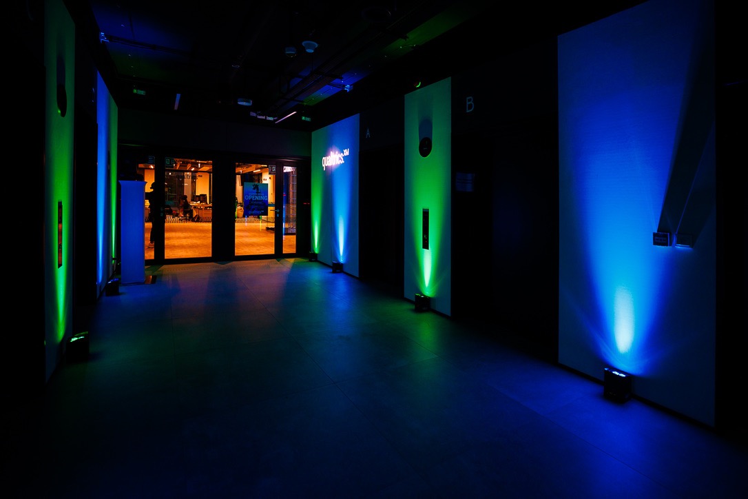 Korytarz z oświetleniem eventowym: białe panele z logo podświetlone niebieskim i zielonym światłem, widoczne osoby w tle za szklanymi drzwiami.