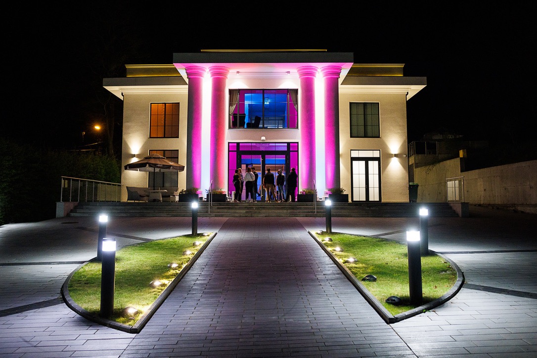 Elegancki budynek z kolumnami oświetlonymi na różowo i biało, przygotowany na imprezę wieczorową, widoczne osoby w wejściu, oświetlona ścieżka prowadząca do budynku.
