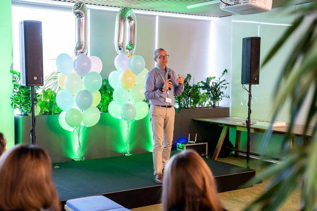 Prelegent z mikrofonem na scenie podczas wydarzenia firmowego, balony w pastelowych kolorach, nagłośnienie po bokach, dekoracje roślinne w tle.