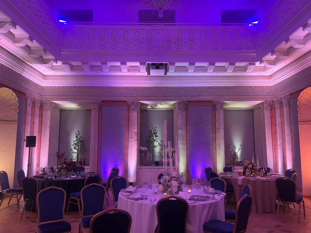 Elegancka sala balowa z okrągłymi stołami, nakryta zastawą i kwiatowymi dekoracjami, oświetlona fioletowym i białym światłem, z kolumnami i sztukaterią.