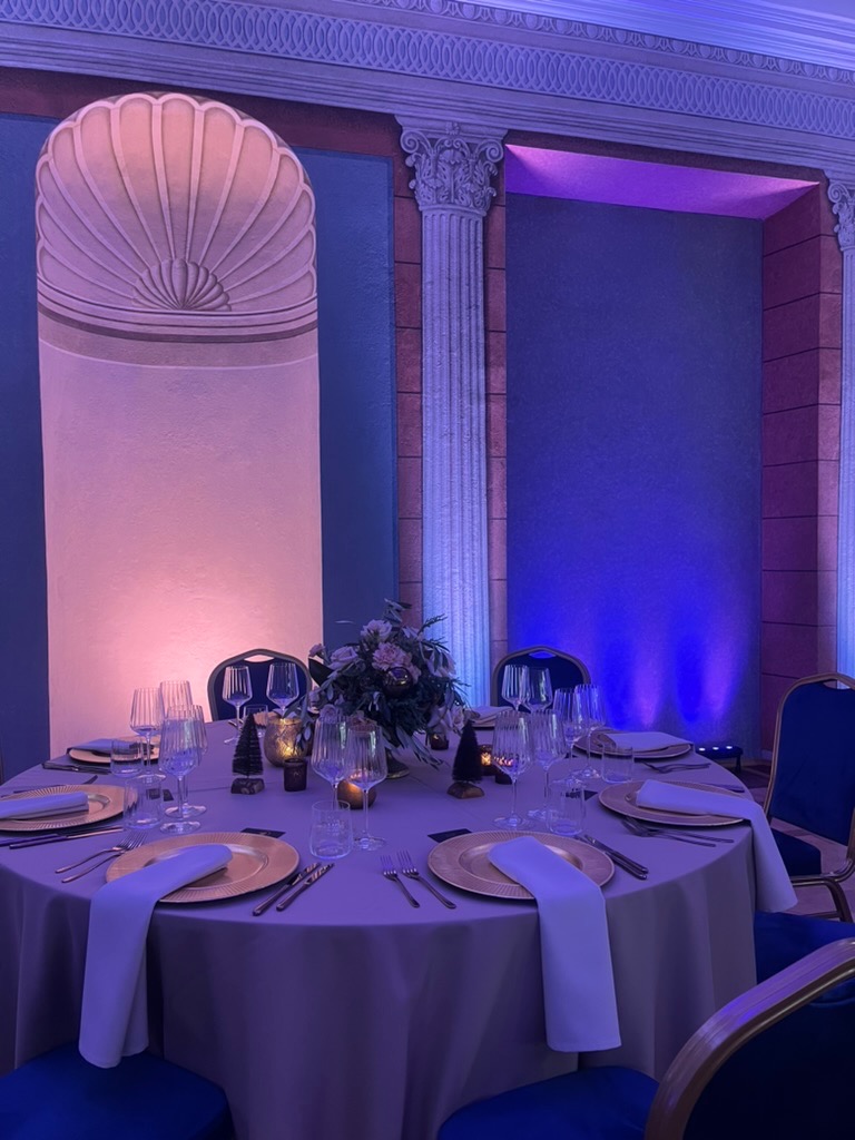 Uroczysty stół z dekoracjami kwiatowymi, złotymi talerzami i białymi serwetkami, otoczony kolumnami i oświetlony fioletowym i różowym światłem.