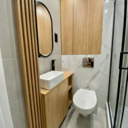 Nowoczesna łazienka z drewnianymi elementami: szafka pod umywalką z pionowymi listwami, owalne lustro w czarnej ramie, wisząca szafka i kabina prysznicowa z czarnymi profilami.