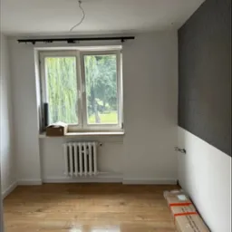 Pusta przestrzeń w trakcie remontu z nową podłogą, grzejnikiem pod oknem i fragmentem ściany z geometryczną tapetą, kable zwisające z sufitu i listwy przypodłogowe w kartonie na podłodze.