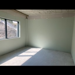 DOM NA MOSORNYM GRONIU Kinga Bryła - Wnętrze pokoju po szpachlowaniu i malowaniu na jasnozielono, z widocznymi przewodami zwisającymi z sufitu i światłem słonecznym wpadającym przez okno.