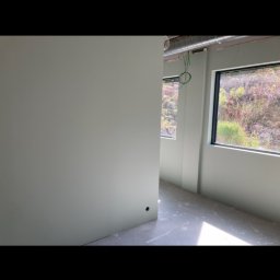 DOM NA MOSORNYM GRONIU Kinga Bryła - Wnętrze w trakcie wykańczania: pomalowane na jasno ściany w odcieniach zieleni, surowy beton na podłodze, widok na zieleń za oknem.