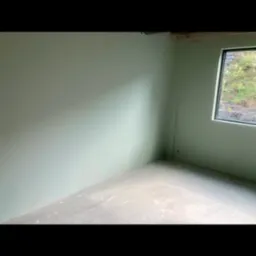 Wnętrze pokoju w trakcie wykańczania: ściany pomalowane na jasnozielony kolor, surowy beton na podłodze, widok za oknem na zieleń.