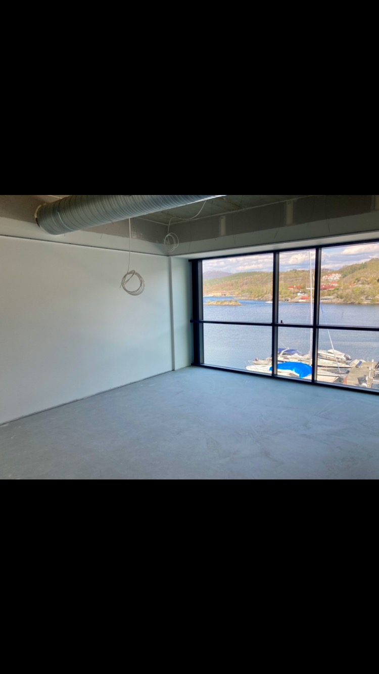 Wnętrze pustego pomieszczenia biurowego z widokiem na zatokę i łodzie przez duże okna; ściany przygotowane do malowania, widoczna wentylacja pod sufitem.