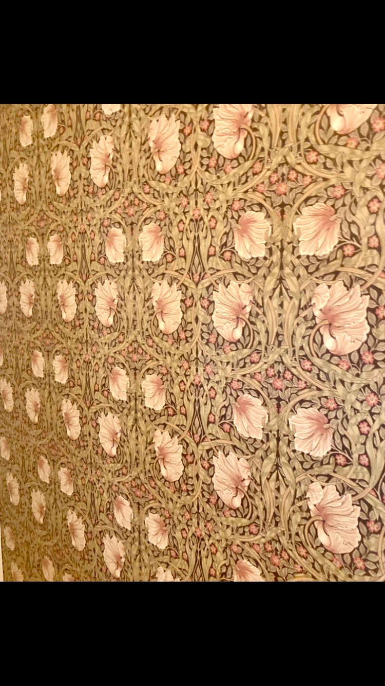 Ściana pokryta tapetą w stylu Morrisa z motywem kwiatów w odcieniach różu i zieleni, widoczne delikatne cieniowanie i faktura tapety.