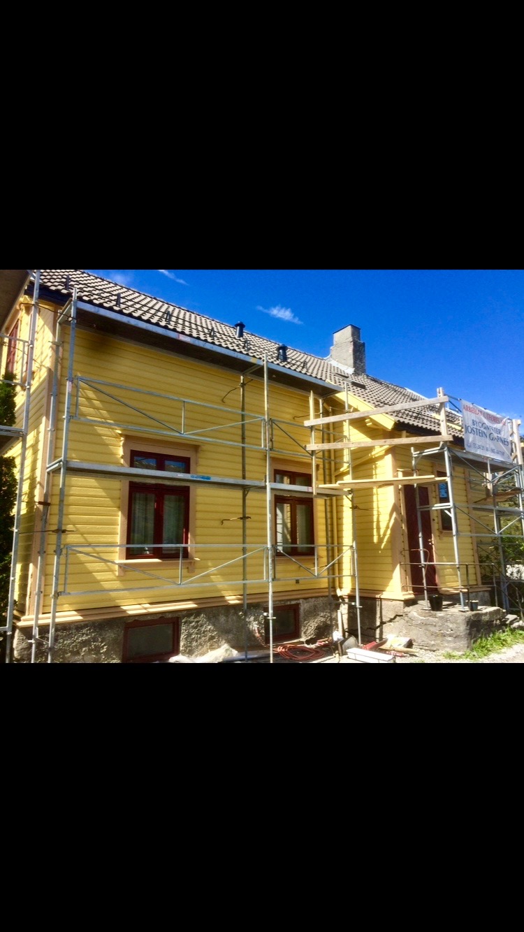 Żółty dom z drewnianą elewacją w trakcie renowacji, otoczony rusztowaniem, z widocznym fragmentem dachu i komina na tle błękitnego nieba.