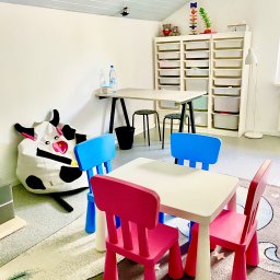 Gabinet Terapii i Rozwoju Dziecka Anna Dajnowska - Jasny pokój terapeutyczny z kolorowymi krzesłami i stolikami dla dzieci, zabawki na półkach, pufa w kształcie krowy. Przytulne i funkcjonalne wnętrze gabinetu logopedy.