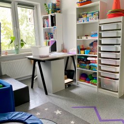 Gabinet Terapii i Rozwoju Dziecka Anna Dajnowska - Jasny gabinet z biurkiem, regałami pełnymi gier i zabawek edukacyjnych, trampoliną i dywanem w gwiazdki. Widoczne pomoce terapeutyczne. Przytulne wnętrze.