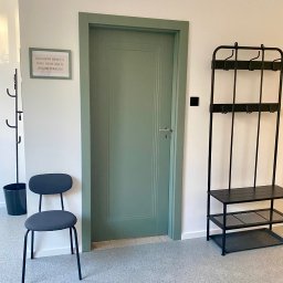 Gabinet Terapii i Rozwoju Dziecka Anna Dajnowska - Korytarz w placówce logopedycznej: zielone drzwi, wieszak na ubrania, krzesło. Na ścianie wisi ramka z informacją dla pacjentów. Jasne, przestronne wnętrze.
