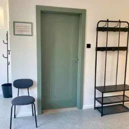 Korytarz w placówce logopedycznej: zielone drzwi, wieszak na ubrania, krzesło. Na ścianie wisi ramka z informacją dla pacjentów. Jasne, przestronne wnętrze.