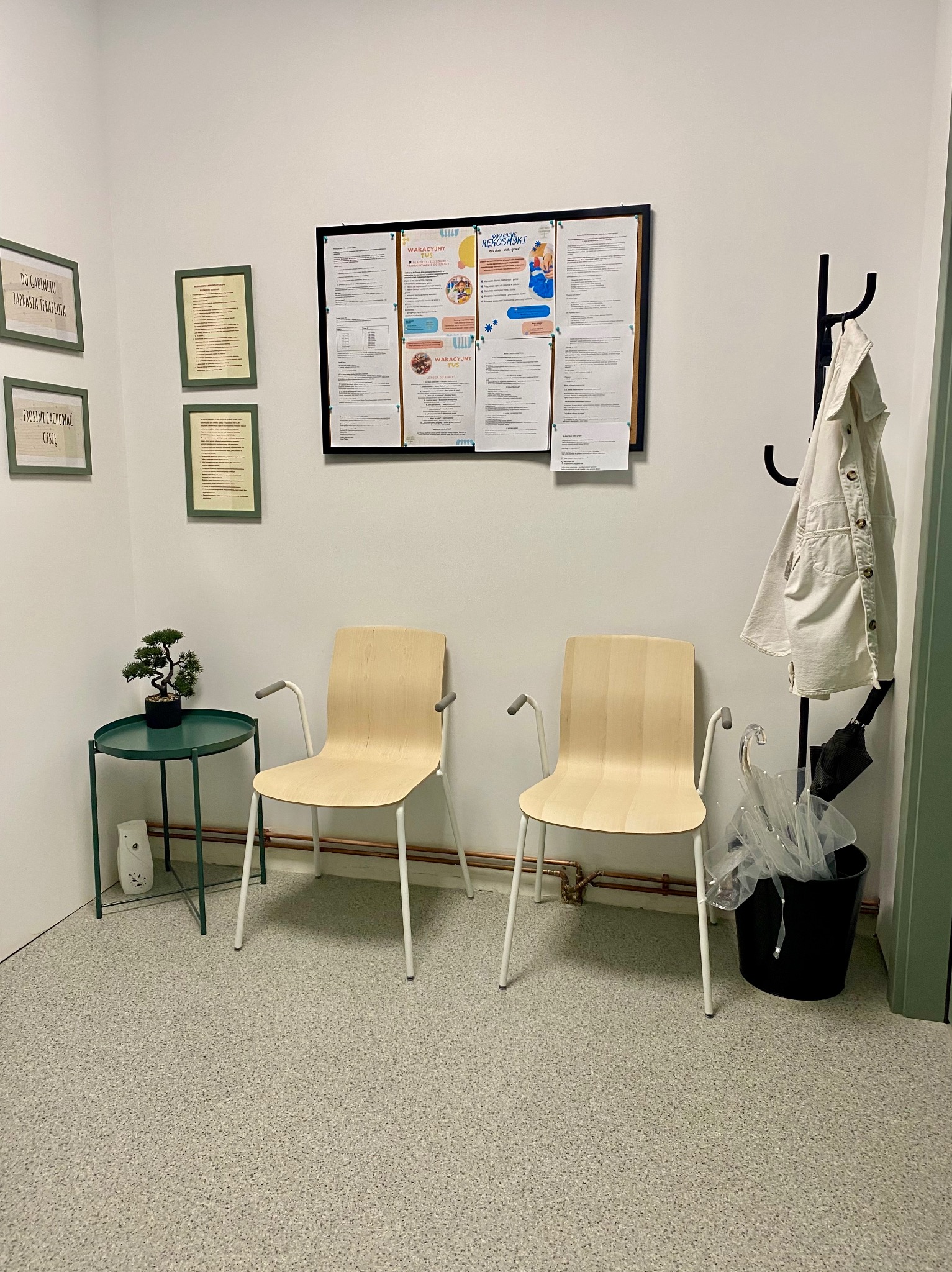 Jasne krzesła w poczekalni gabinetu z informacjami na ścianie, stolik z bonsai, wieszak z płaszczem i parasolem. Przytulne, minimalistyczne wnętrze.