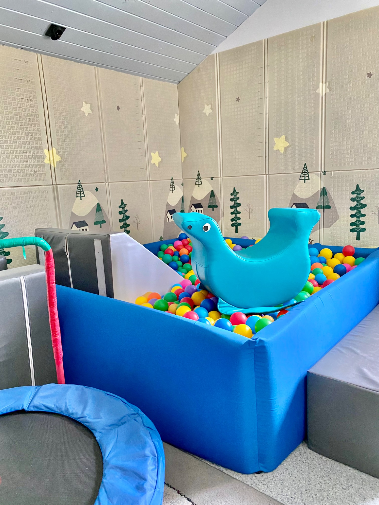 Kącik zabaw dla dzieci z basenem z kulkami, trampoliną i miękkimi elementami w stonowanej kolorystyce. Ściany wyłożone matami z motywem gór i drzew.