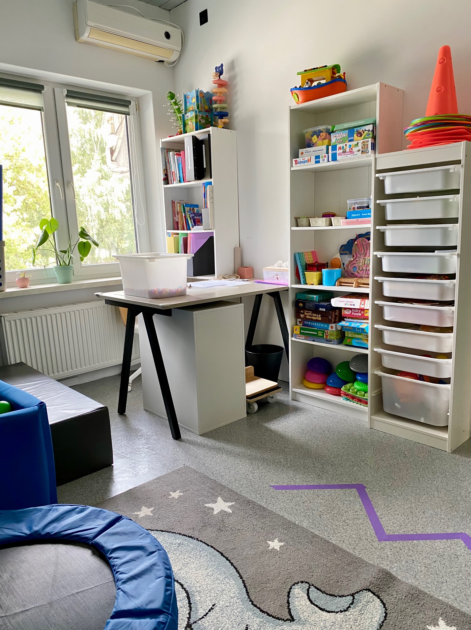 Jasny gabinet z biurkiem, regałami pełnymi gier i zabawek edukacyjnych, trampoliną i dywanem w gwiazdki. Widoczne pomoce terapeutyczne. Przytulne wnętrze.
