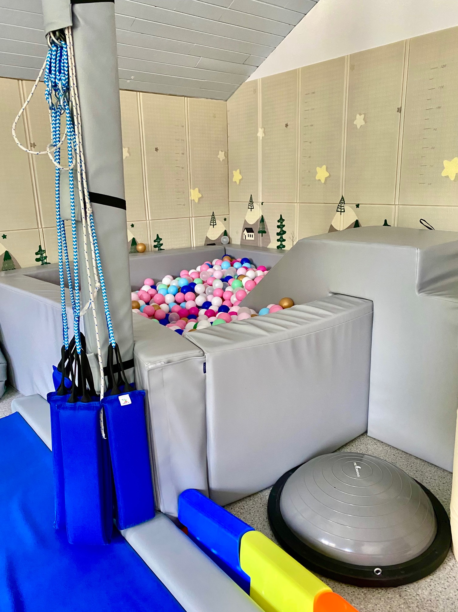 Sala do terapii integracji sensorycznej: suchy basen z piłeczkami, podwieszane platformy, mata podłogowa z motywem gór i gwiazd, półkula do ćwiczeń równowagi.