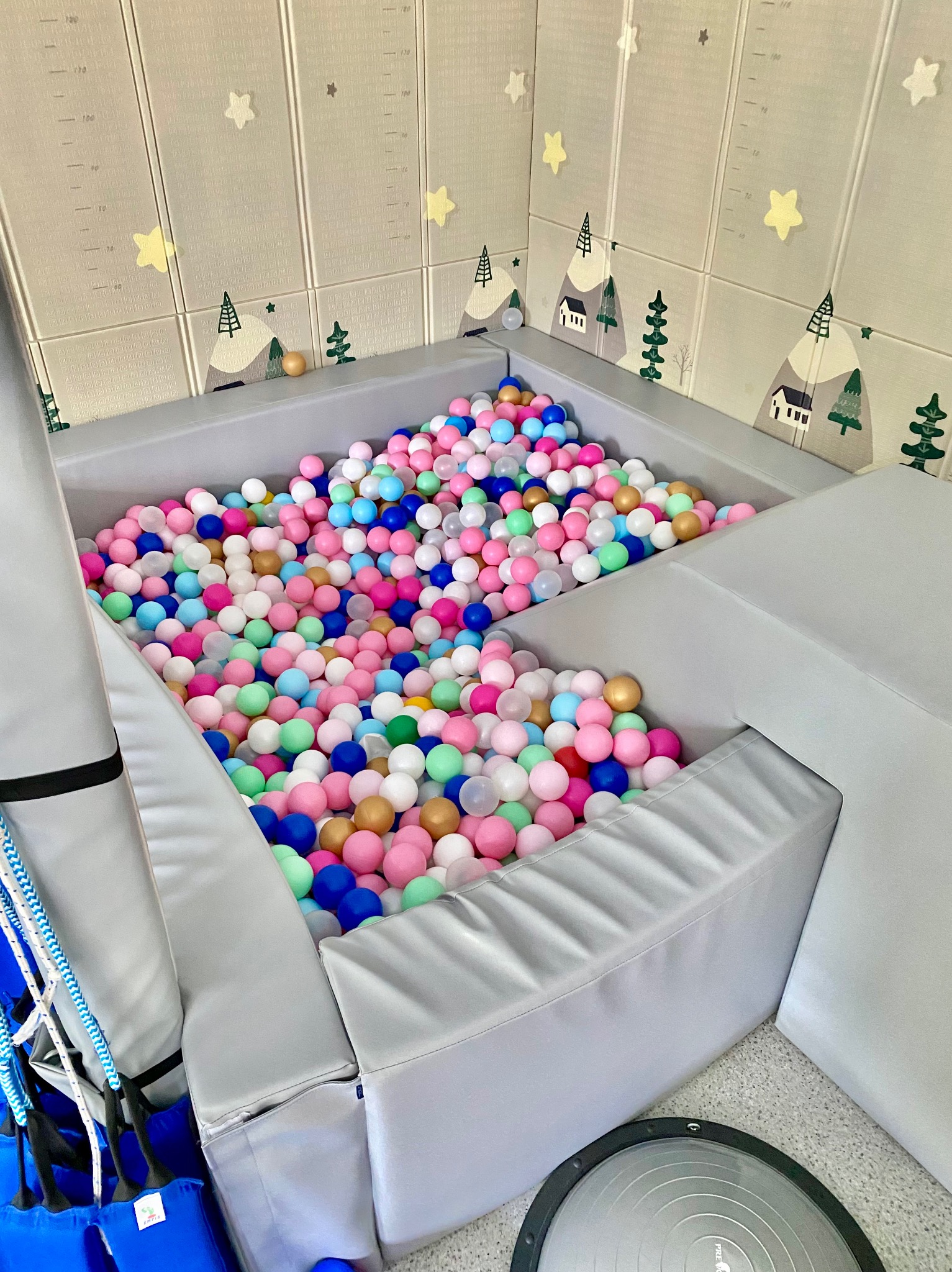 Kolorowy basen z piłeczkami w gabinecie logopedycznym, otoczony ścianami z motywem gór i drzew. Widoczny sprzęt rehabilitacyjny.