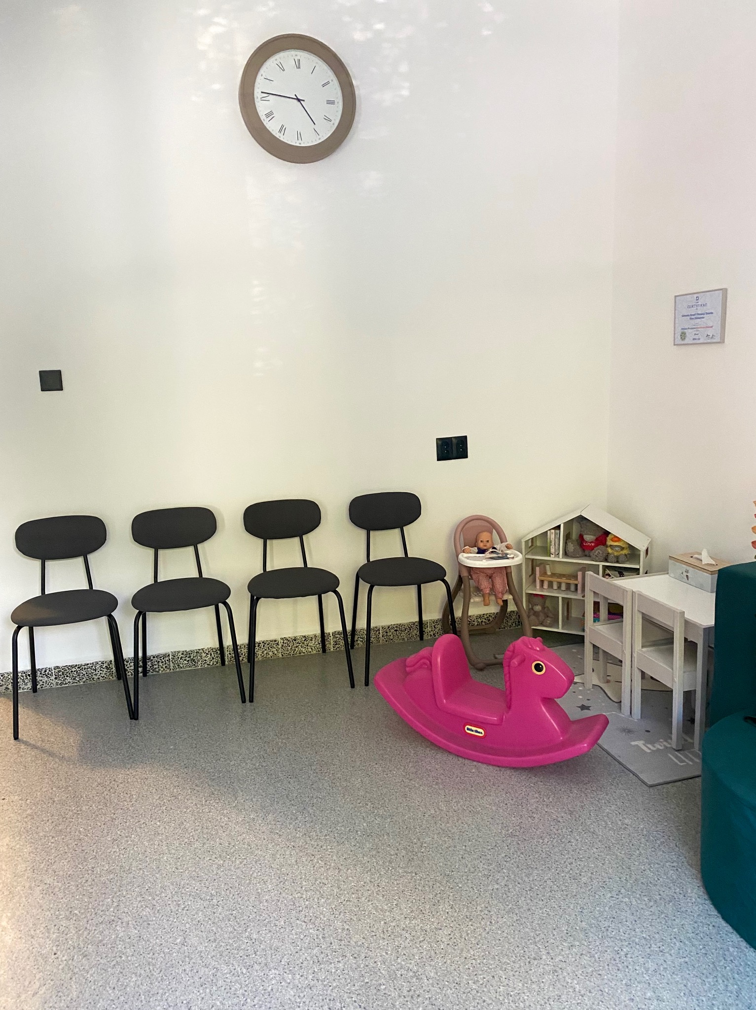 Poczekalnia: cztery krzesła, zabawki (konik, domek dla lalek), zegar na ścianie, certyfikat. Przyjazna przestrzeń dla dzieci.