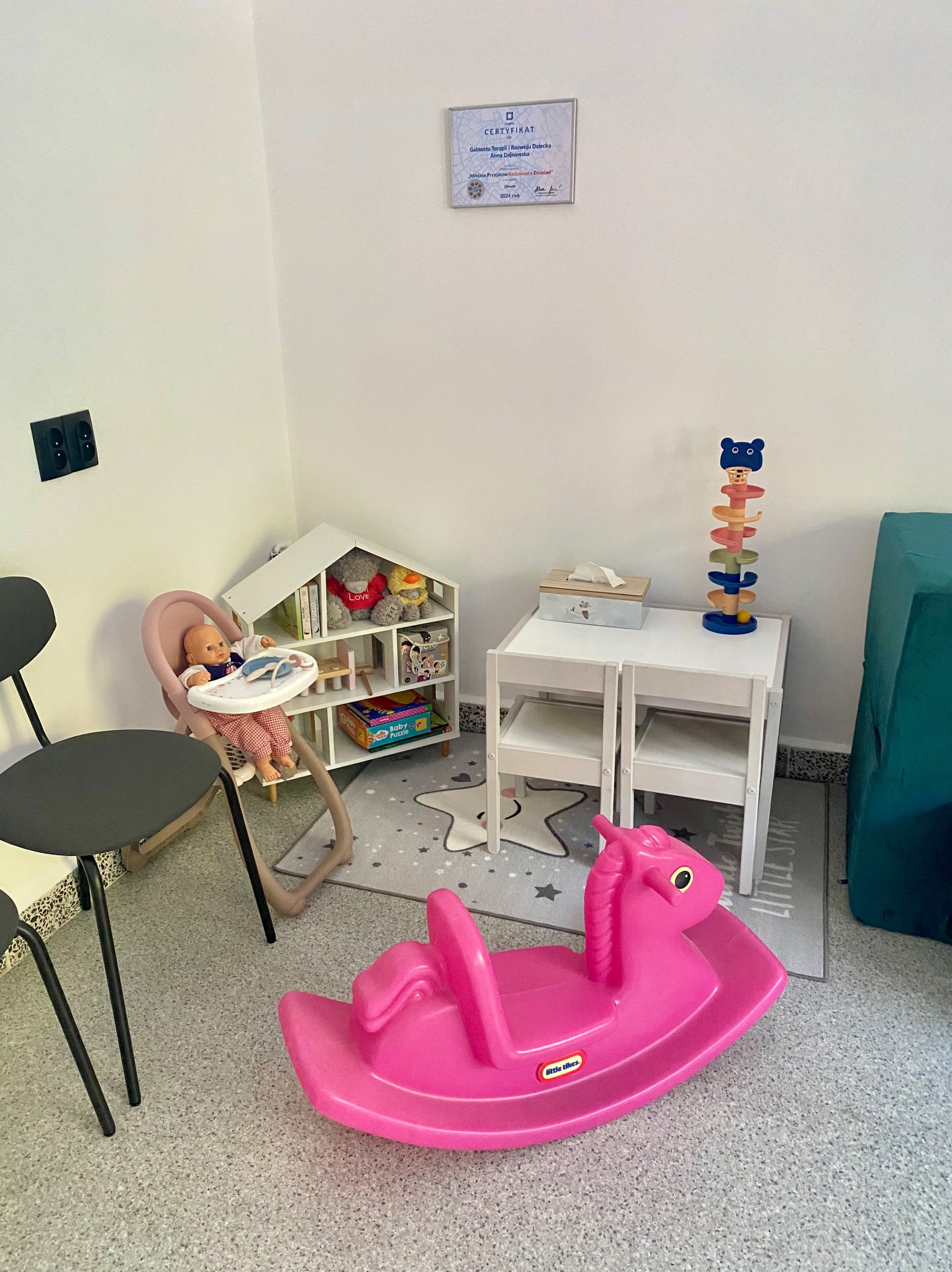 Jasny gabinet z certyfikatem na ścianie, stolikiem, krzesłami, półką z zabawkami, lalką w krzesełku i różowym konikiem na biegunach, tworzący przyjazną przestrzeń dla dzieci.