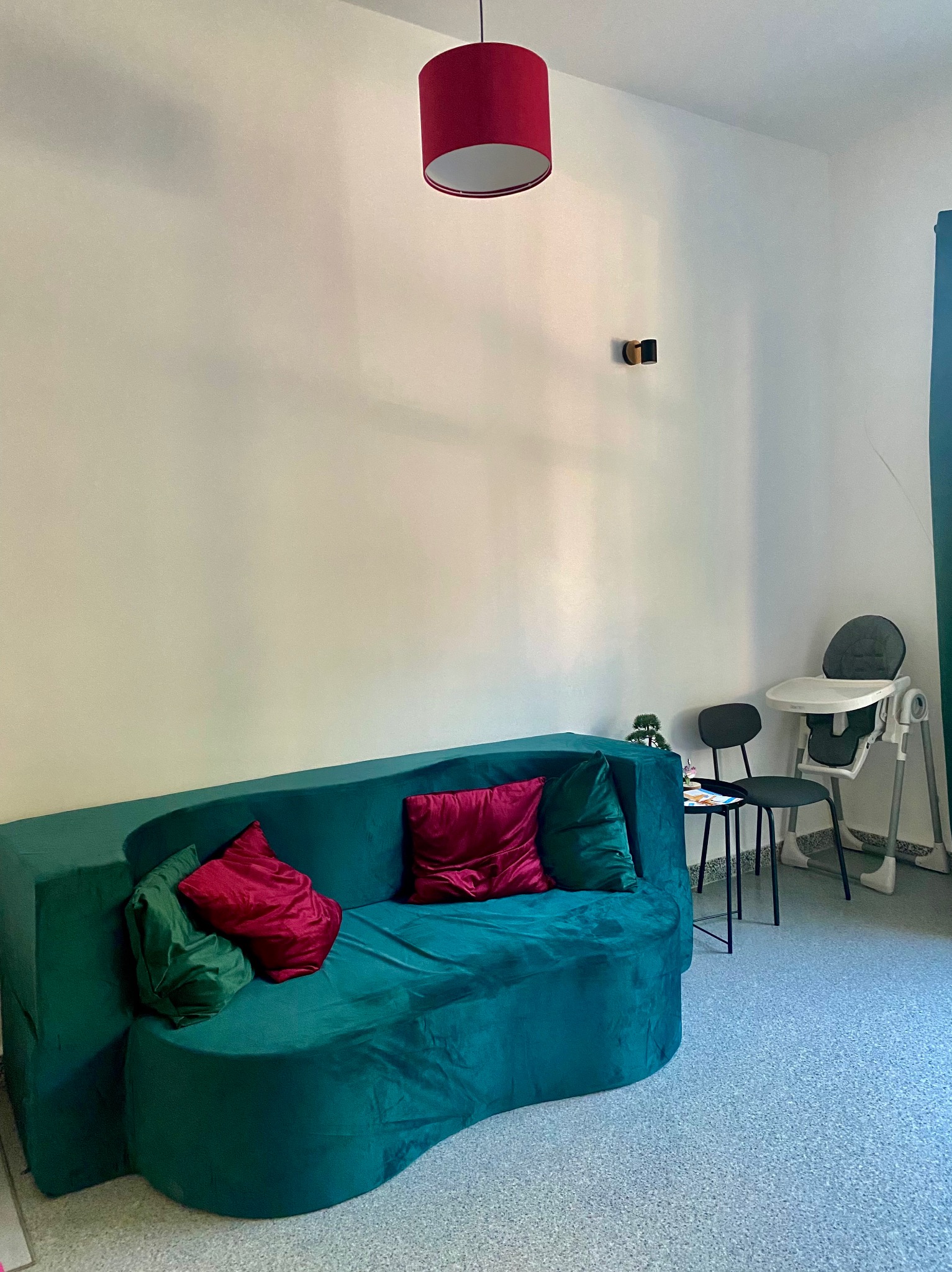 Kącik terapeutyczny: zielona sofa z poduszkami, stolik z dekoracją, krzesło i krzesełko do karmienia. Czerwona lampa i jasne ściany tworzą przyjazną atmosferę.