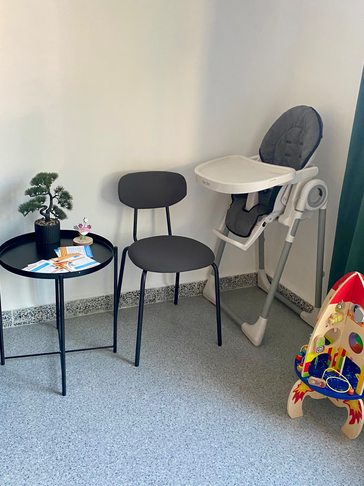 Przytulny kącik dla dzieci z krzesłem do karmienia, stolikiem z bonsai i drewnianą zabawką rakietą w gabinecie, prawdopodobnie logopedycznym.