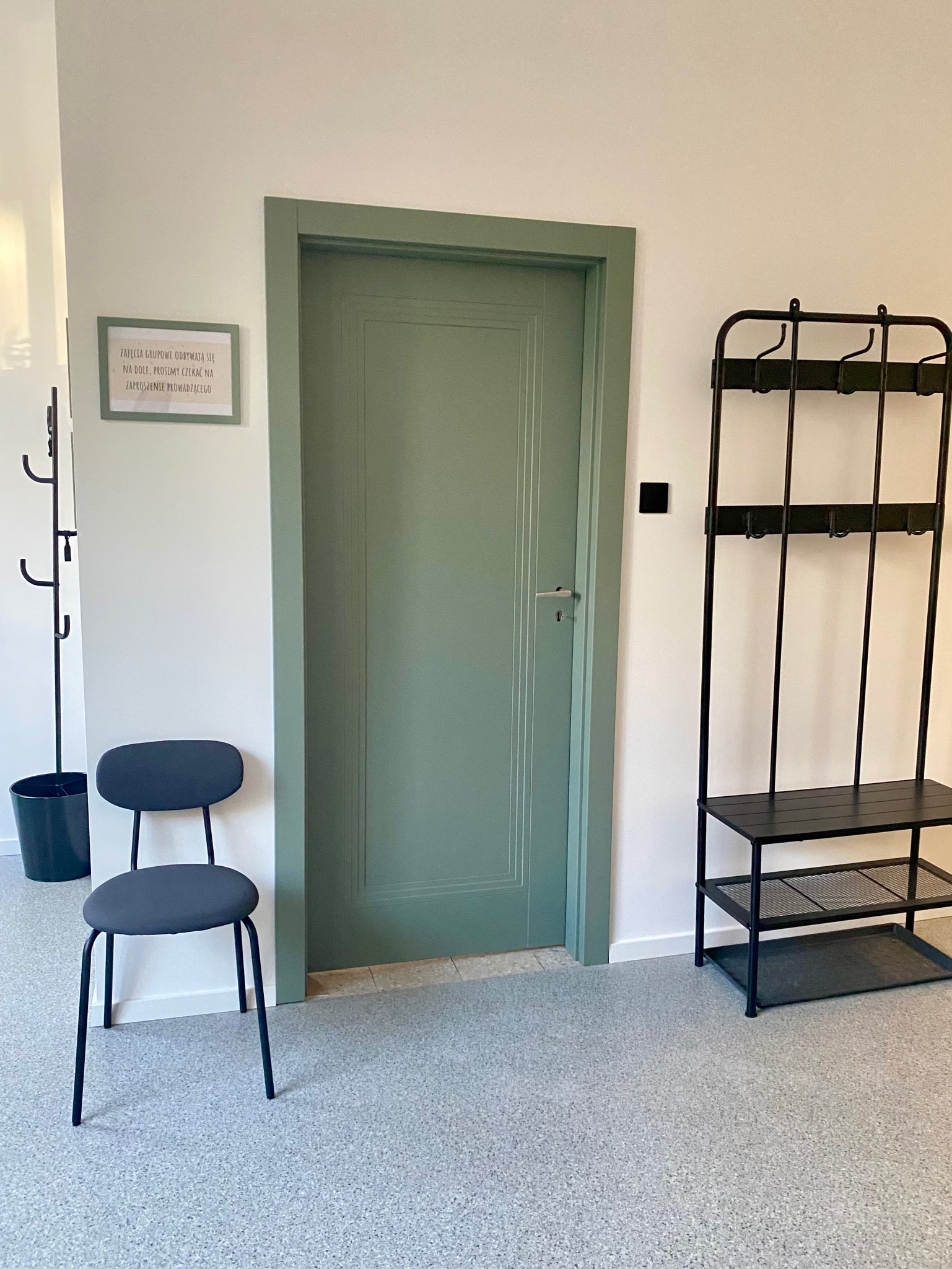 Korytarz w placówce logopedycznej: zielone drzwi, wieszak na ubrania, krzesło. Na ścianie wisi ramka z informacją dla pacjentów. Jasne, przestronne wnętrze.