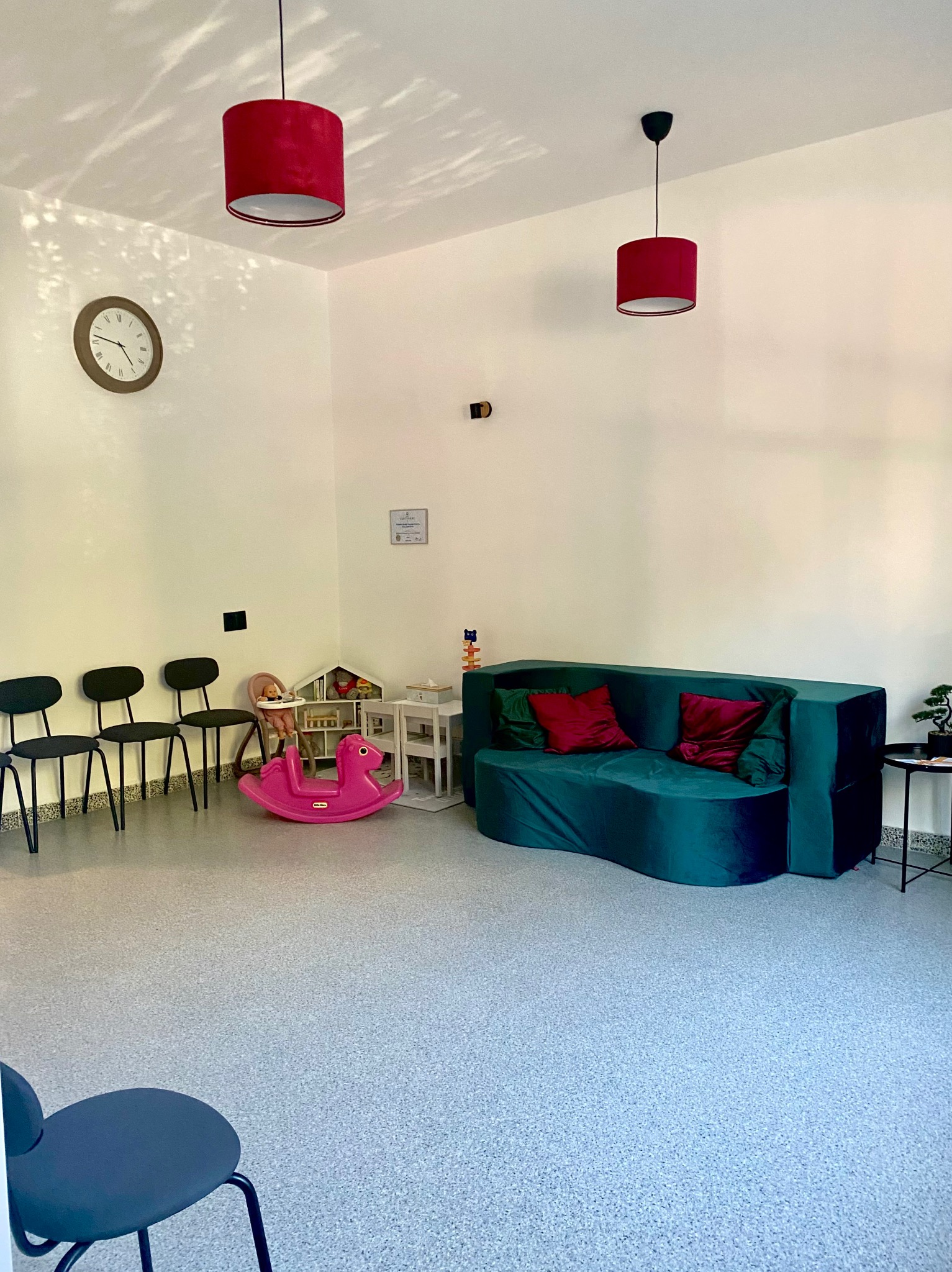 Jasna poczekalnia logopedy z zieloną sofą, różowym konikiem na biegunach, zegarem i czerwonymi lampami. Przytulne wnętrze dla dzieci.