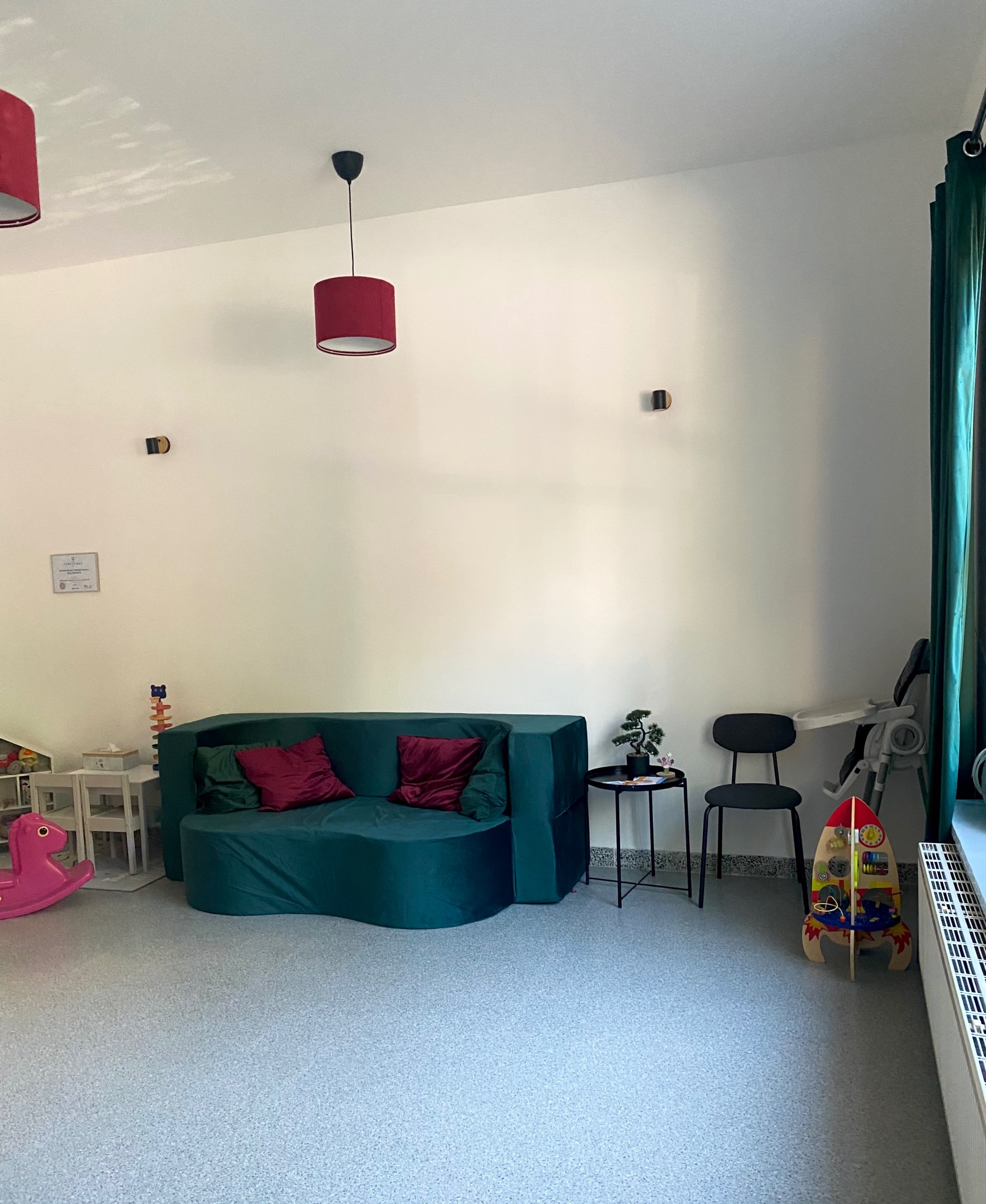 Kącik terapeutyczny dla dzieci: zielona sofa z bordowymi poduszkami, zabawki, krzesło i stolik. Jasne wnętrze z elementami dekoracyjnymi.