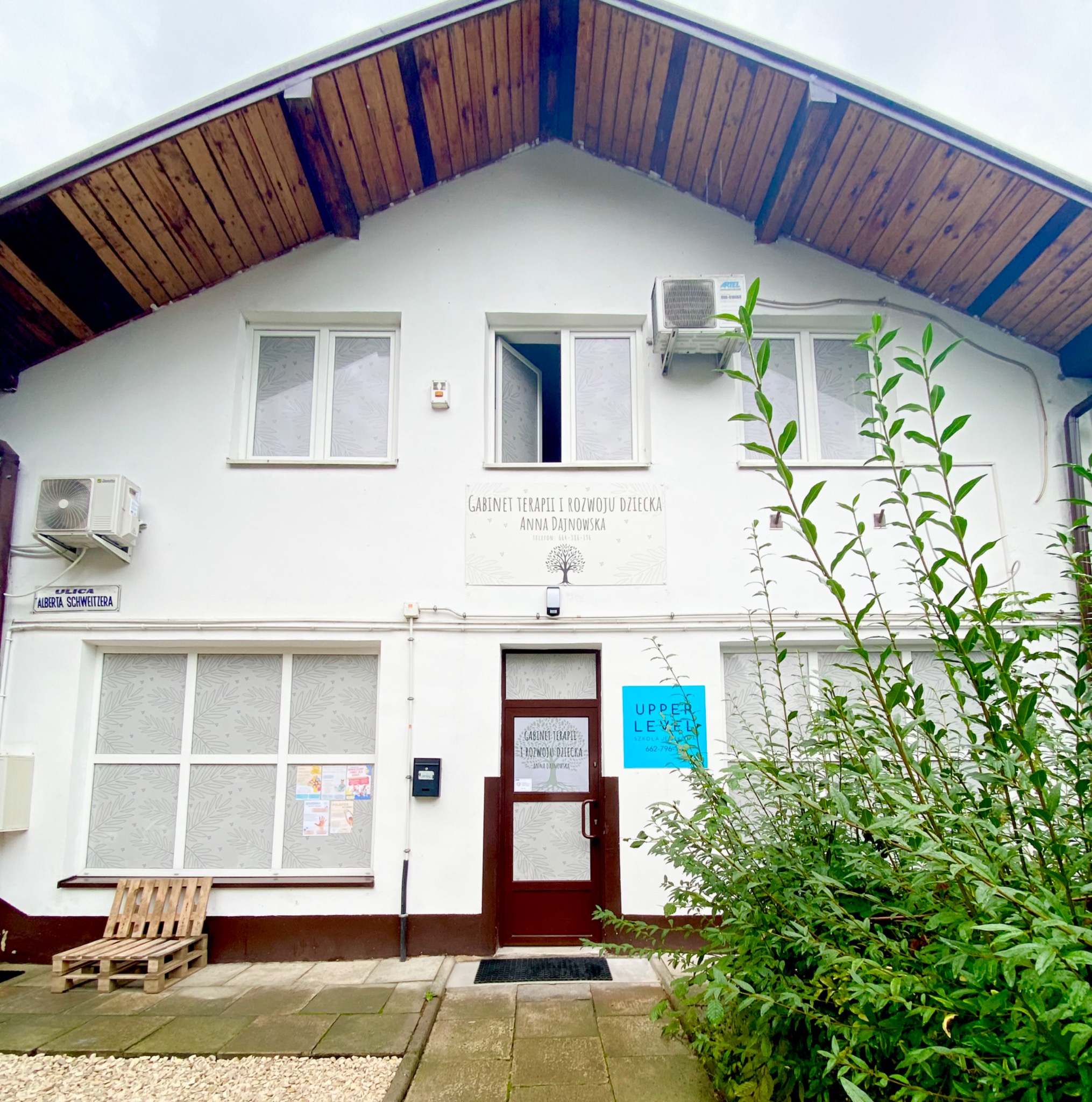 Gabinet terapii i rozwoju dziecka z nazwą na fasadzie, brązowe drzwi wejściowe, okna z naklejkami i krzesło z palet na zewnątrz.