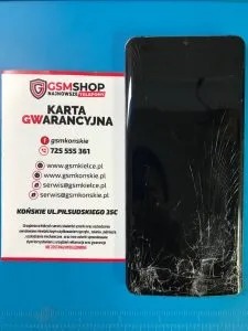Uszkodzony ekran smartfona z widocznymi pęknięciami, leżący obok karty gwarancyjnej GSMshop na niebieskim podłożu. Widoczny adres i numer telefonu serwisu.
