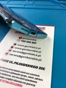 Uszkodzony smartfon z widocznymi śladami upadku leżący na karcie wizytowej serwisu GSM z danymi kontaktowymi i adresem. Wizytówka na niebieskiej macie serwisowej.