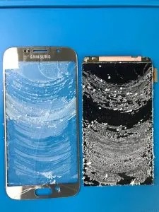 Uszkodzony telefon Samsung z odseparowanym, pękniętym ekranem dotykowym i wyświetlaczem, leżący na niebieskim tle. Widoczne ślady kleju i uszkodzenia mechaniczne.