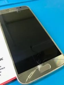 Uszkodzony smartfon Samsung Galaxy S6 z pękniętym ekranem u dołu, leżący na niebieskiej macie serwisowej obok opakowania części zamiennej.