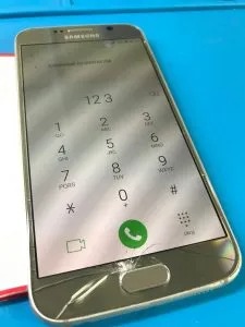 Uszkodzony smartfon Samsung z pękniętym ekranem, na którym wyświetlona jest klawiatura numeryczna. Widoczne ślady użytkowania i zabrudzenia na obudowie oraz ekranie. Telefon leży na niebieskim...