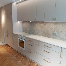 AGStolarstwo - Jasna kuchnia z drewnianą podłogą w jodełkę, beżowe fronty szafek z poziomymi uchwytami, piekarnik w zabudowie, dekoracyjna płytka nad blatem, oświetlenie LED podszafkowe.