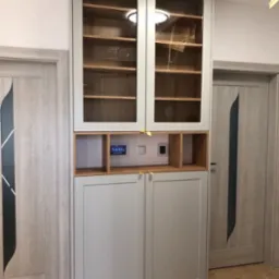 Wysoka, szara szafa z przeszklonymi górnymi drzwiczkami i drewnianymi półkami wewnątrz, zintegrowana z panelem sterowania domem i oświetleniem LED, umieszczona między dwiema szarymi drzwiami.