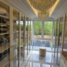 Elegancka garderoba z przeszklonymi szafami, złotymi uchwytami i kryształowym żyrandolem. Widok na zieleń za oknem.